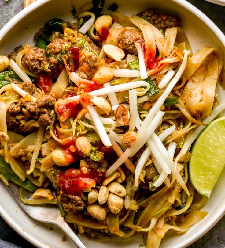 A bowl of ground beef pad thai topped with peanuts, bean sprouts, and lime wedges, with a fork on the side.