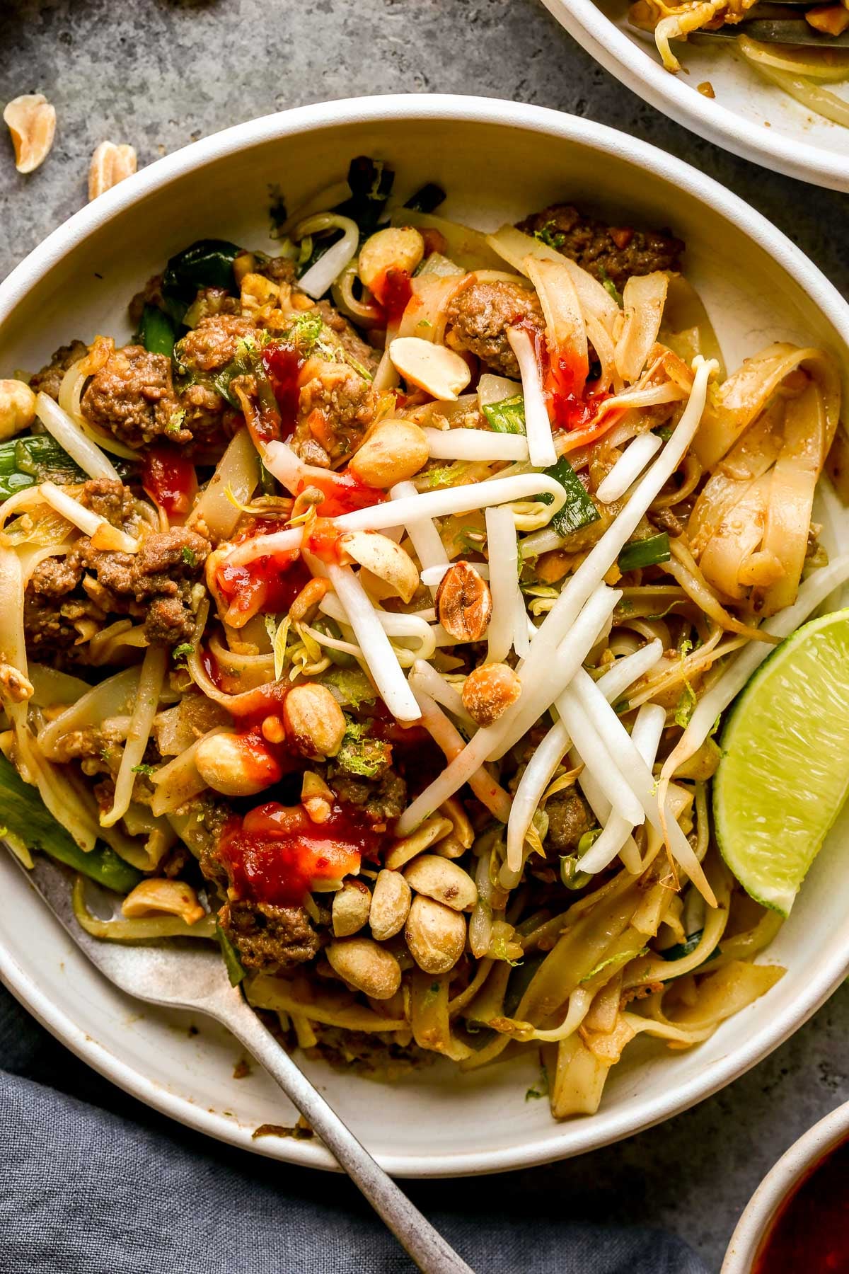 A bowl of ground beef pad thai topped with peanuts, bean sprouts, and lime wedges, with a fork on the side.