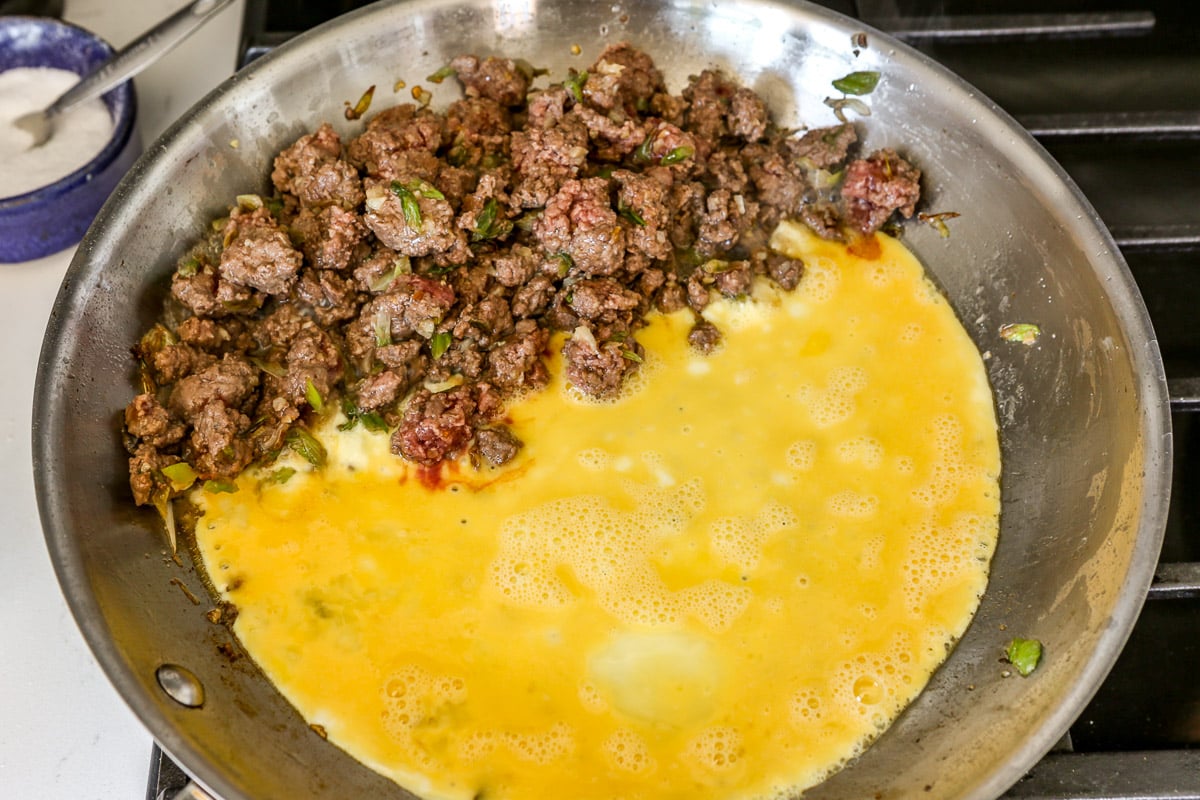 Browned ground beef cooking in a in a large stainless steel skillet one one side and scrambled eggs cooking on the other side.