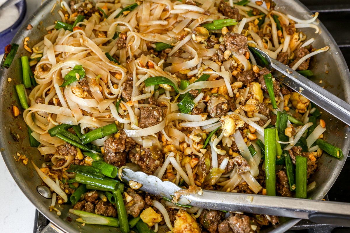 A pan of beef stir-fry with rice noodles, green onions, bean sprouts, peanuts, and metal tongs set in the pan.