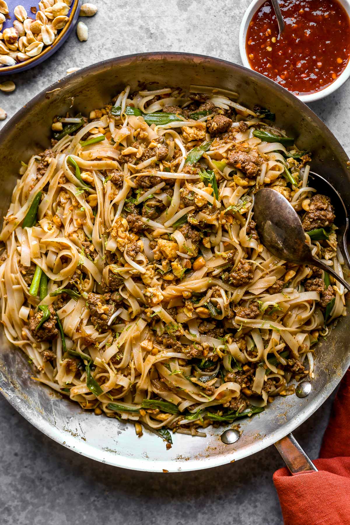 A skillet filled with stir-fried noodles, ground beef, and vegetables, with a spoon and bowls of sauce and peanuts nearby.