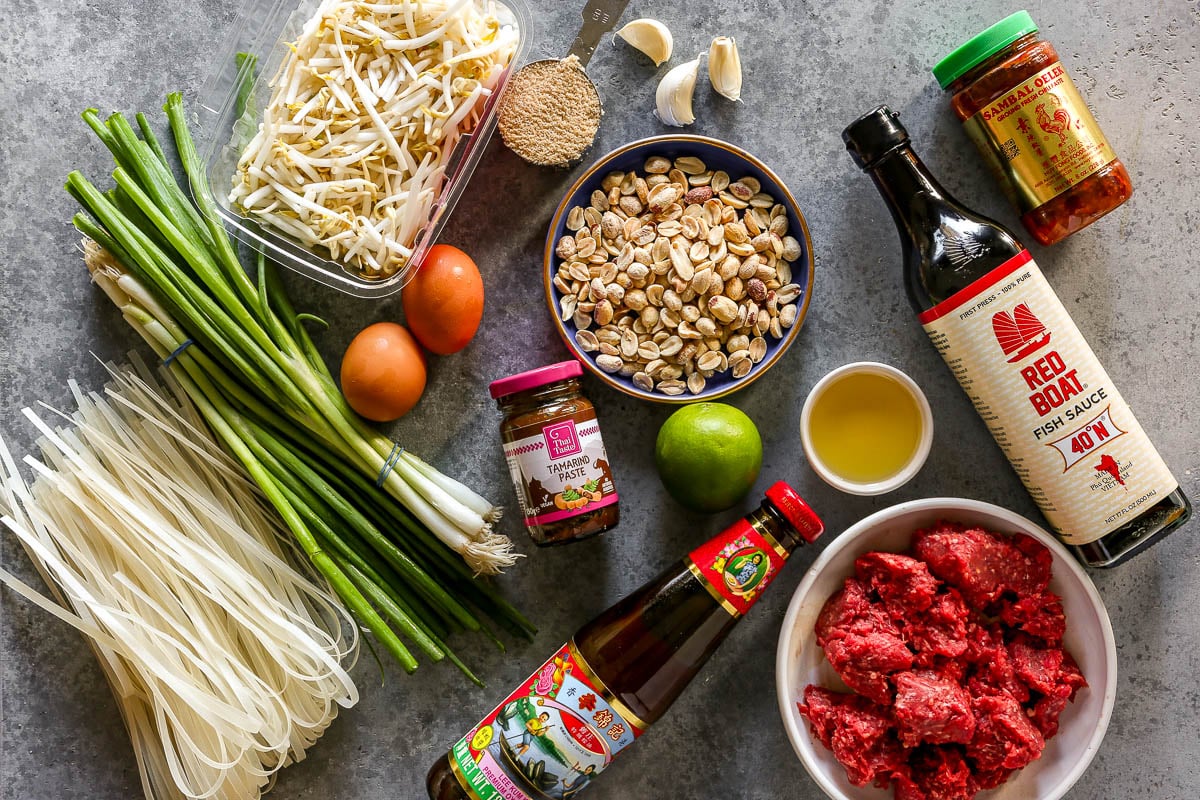 Ingredients for beef and noodles set out on a counter including rice noodles, scallions, mung bean sprouts, eggs, garlic, brown sugaar, peanuts, tamarind paste, oyster sauce, lime, oil, ground beef, fish sauce and hot sauce.