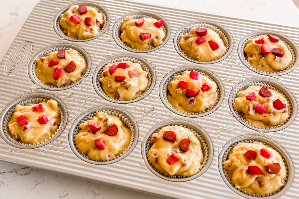 Muffin batter in paper liners in a muffin tin, topped with pieces of diced rhubarb.