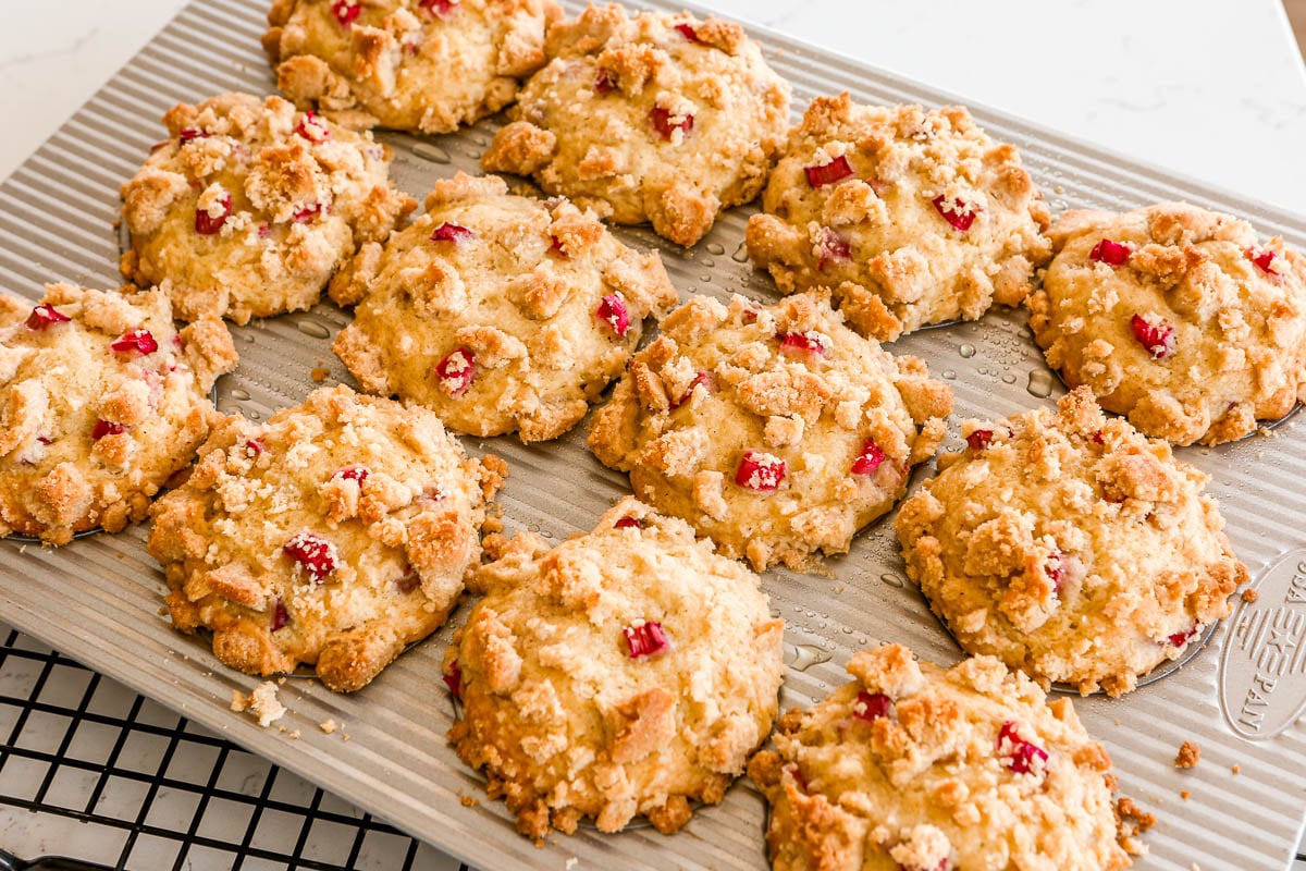 Baked rhubarb muffins with streusel in a silver muffin tin set on a black wire rack on a white counter.
