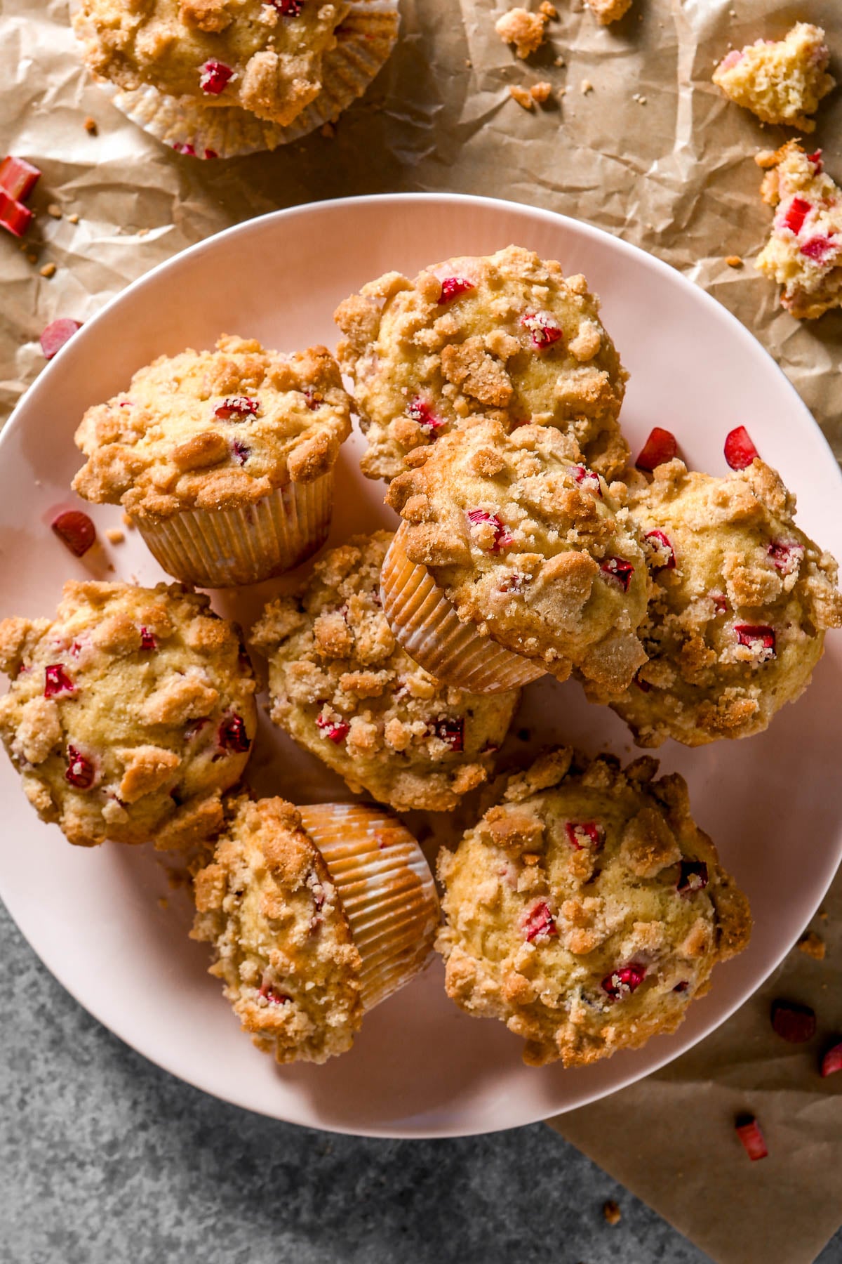 A plate of crumb-topped rhubarb muffins with bits of rhubarb, surrounded by crumbs on parchment paper.