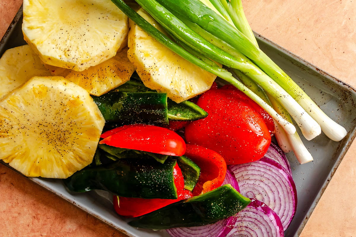 Pineapple, red peppers, green peppers, green onions, and red onions arranged on a baking sheet.