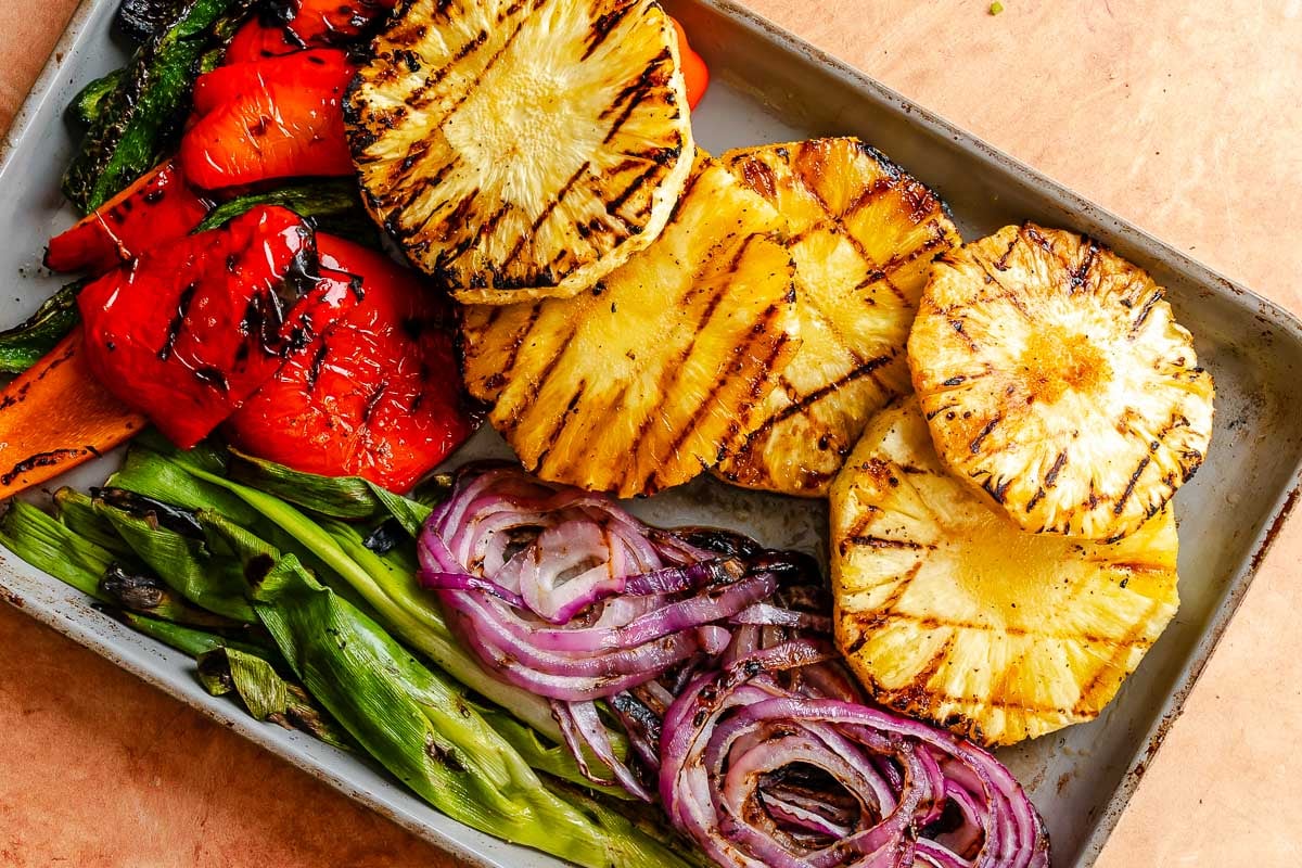 Grilled pineapple, red peppers, green peppers, green onions, and red onions arranged on a baking sheet.