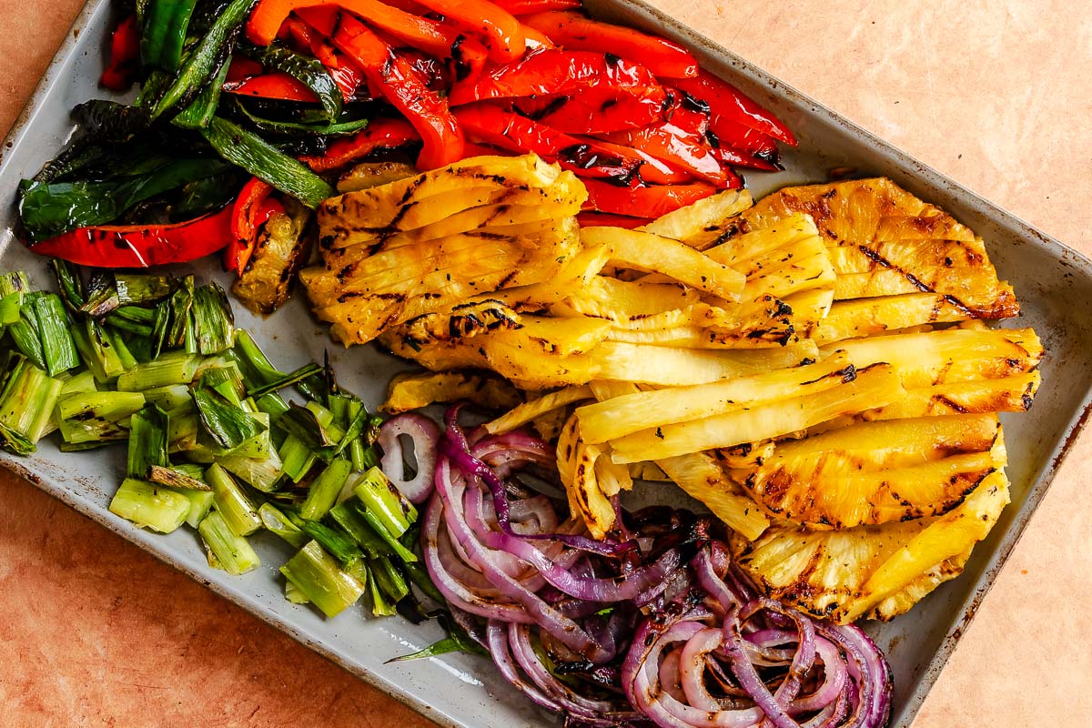 Sliced grilled pineapple, red peppers, green peppers, green onions, and red onions arranged on a baking sheet.
