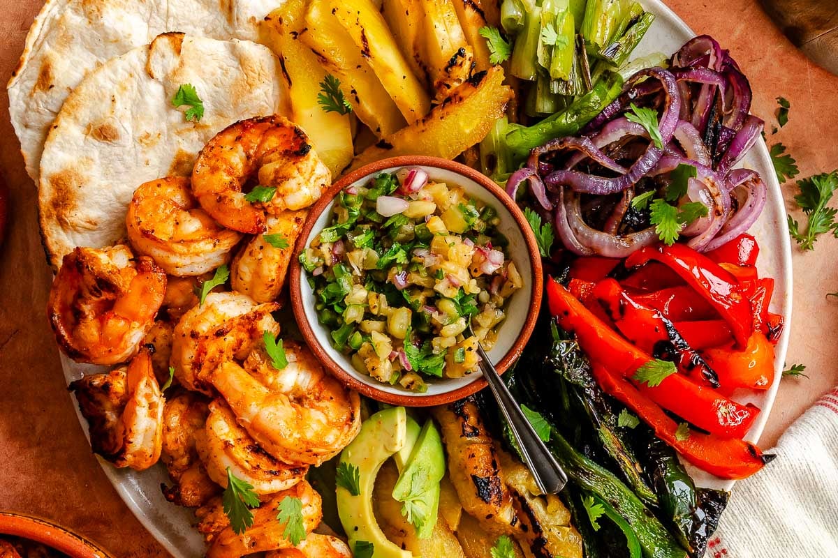 A platter with grilled shrimp fajitas featuring pineapple, avocado, grilled veggies, salsa, and tortillas on the side.