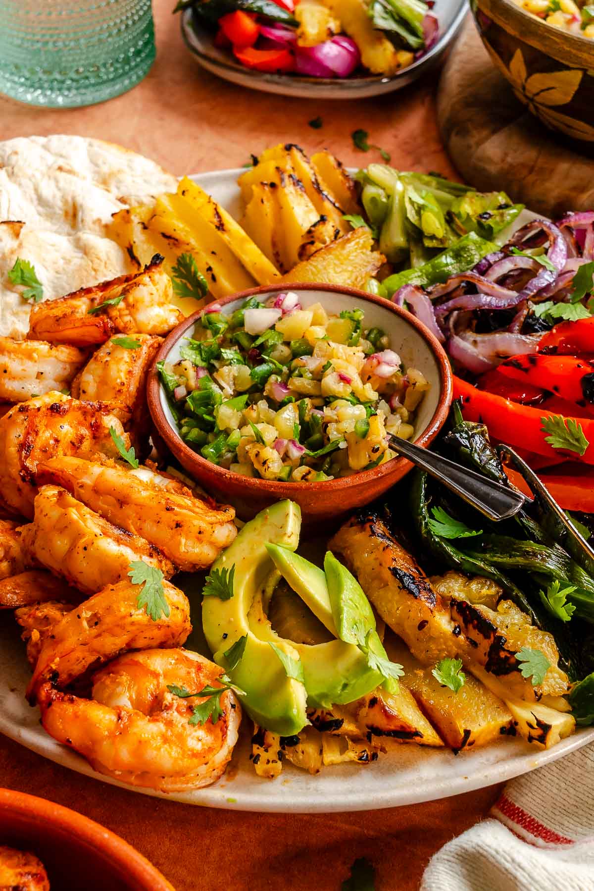 A platter with grilled shrimp fajitas featuring pineapple, avocado, grilled veggies, salsa, and tortillas on the side.