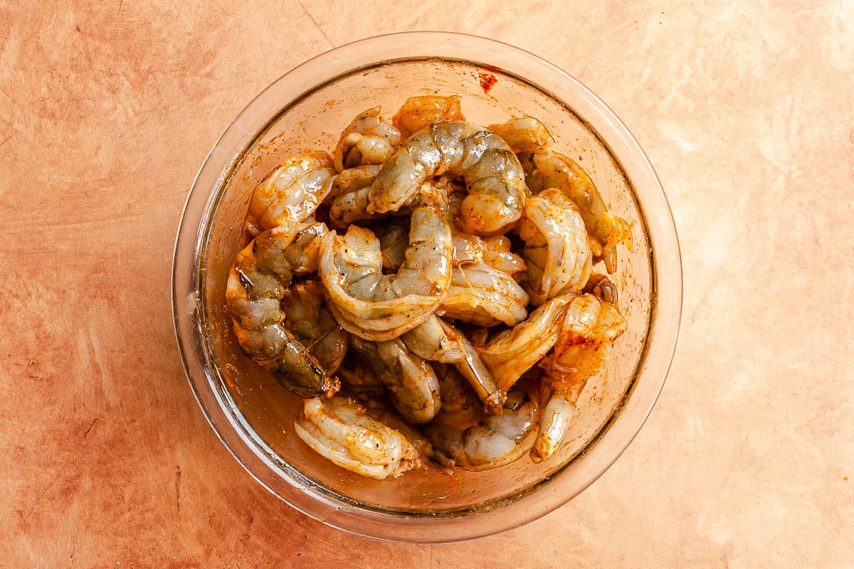 Raw shrimp marinating in a glass bowl on a light brown surface.