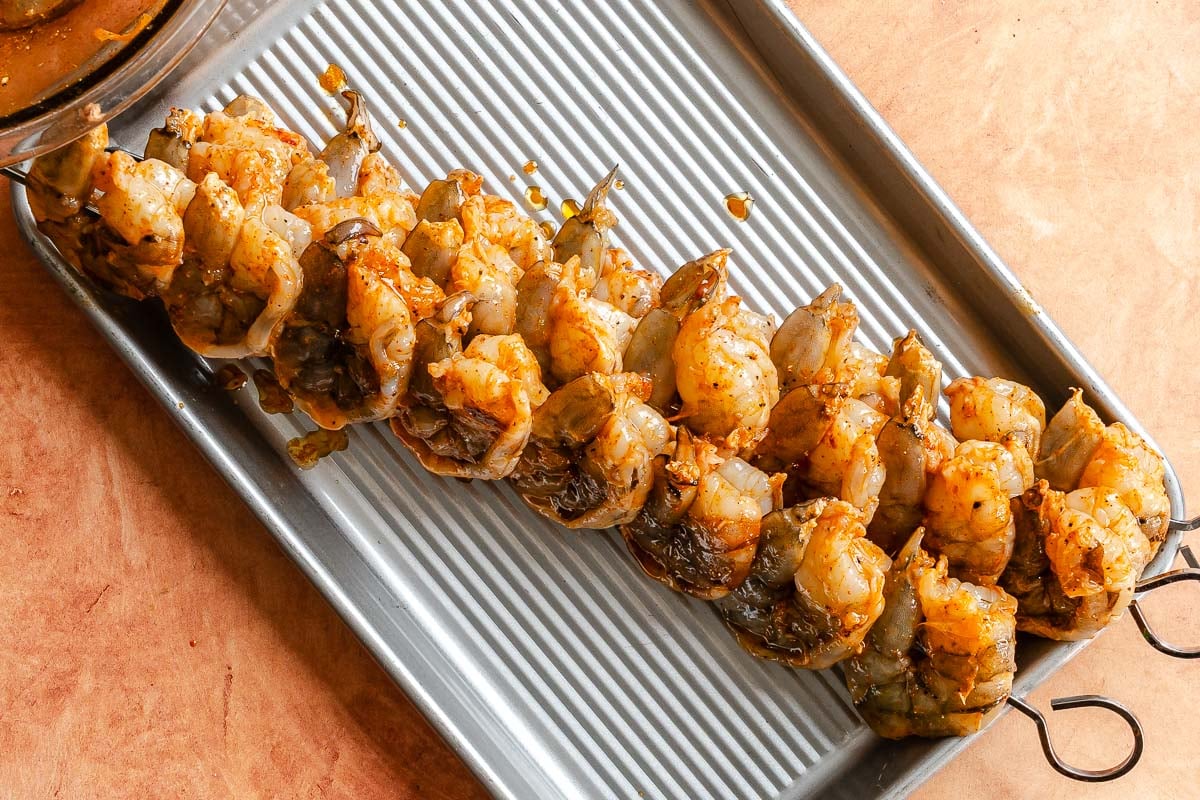 Two metal skewers with marinated shrimp are placed on a silver baking tray.