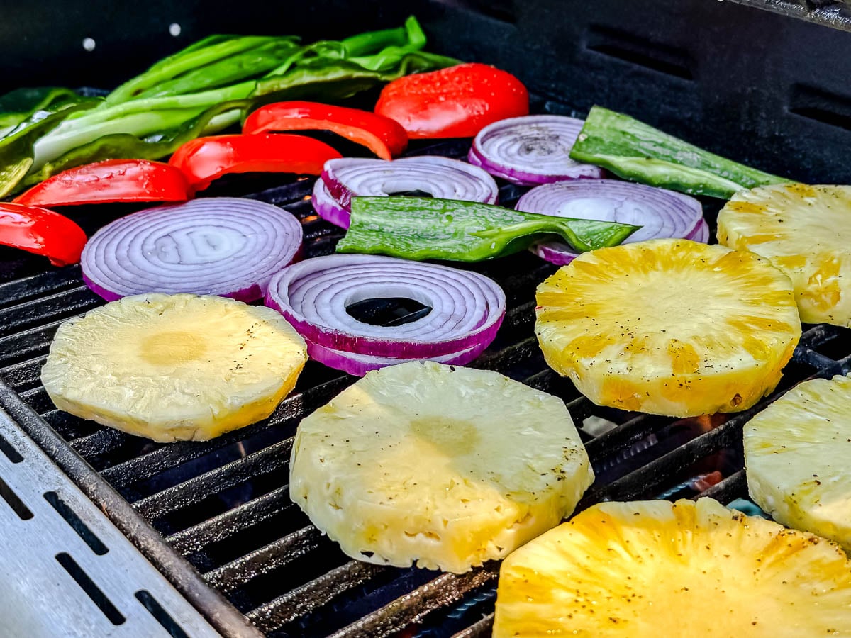 Slices of pineapple, red onion, red bell pepper, poblano pepper, and green onions grilling on a barbecue.