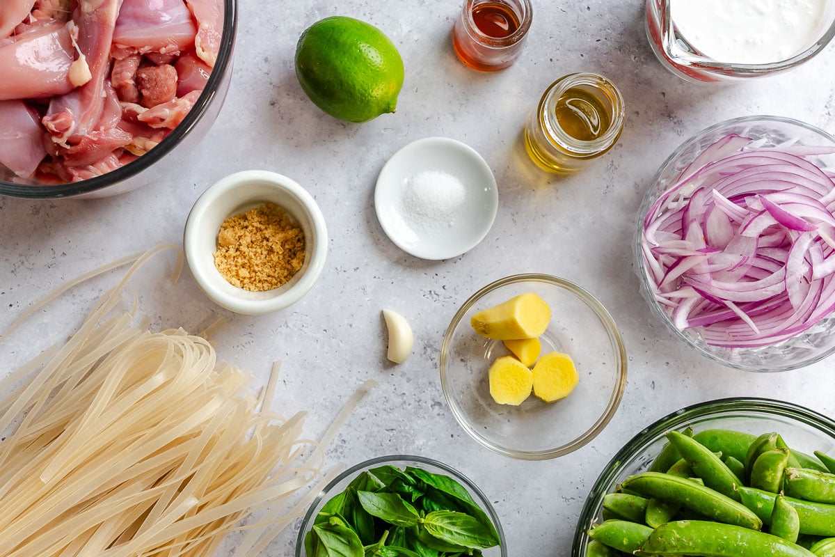 Raw chicken, dry rice noodles, sliced onions, lime, garlic, ginger, snap peas, basil, coconut milk, salt and oil set out on a counter.