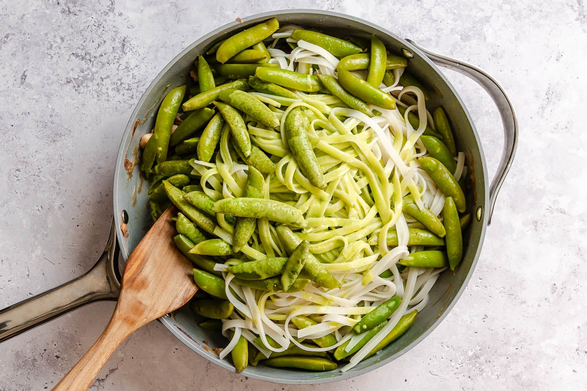 A skillet with snap peas, rice noodles, and a green sauce poured over top; stirred with a wooden spatula on a light countertop.