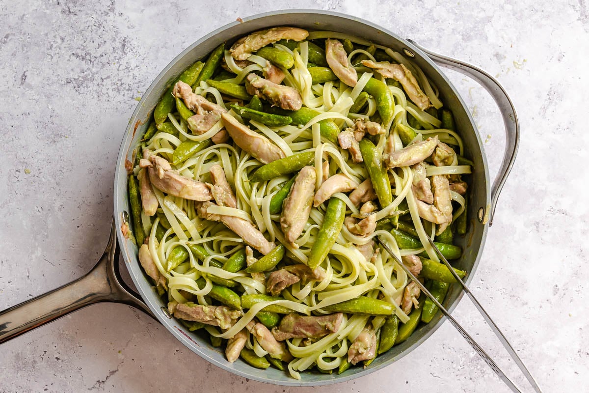Large skillet with rice noodles, cooked chicken thighs, and snap peas all tossed in green coconut sauce. Tongs rest inside. Fresh herbs and ginger nearby.