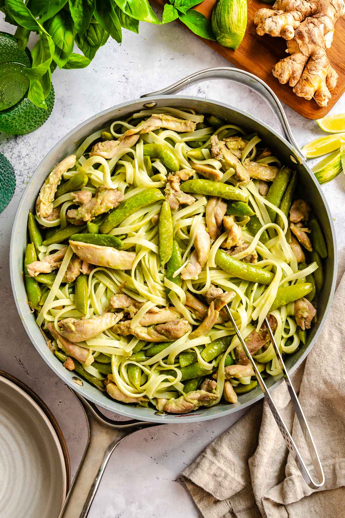 Large skillet with pasta, chicken strips, and snap peas. Tongs rest inside. Fresh herbs and ginger nearby.