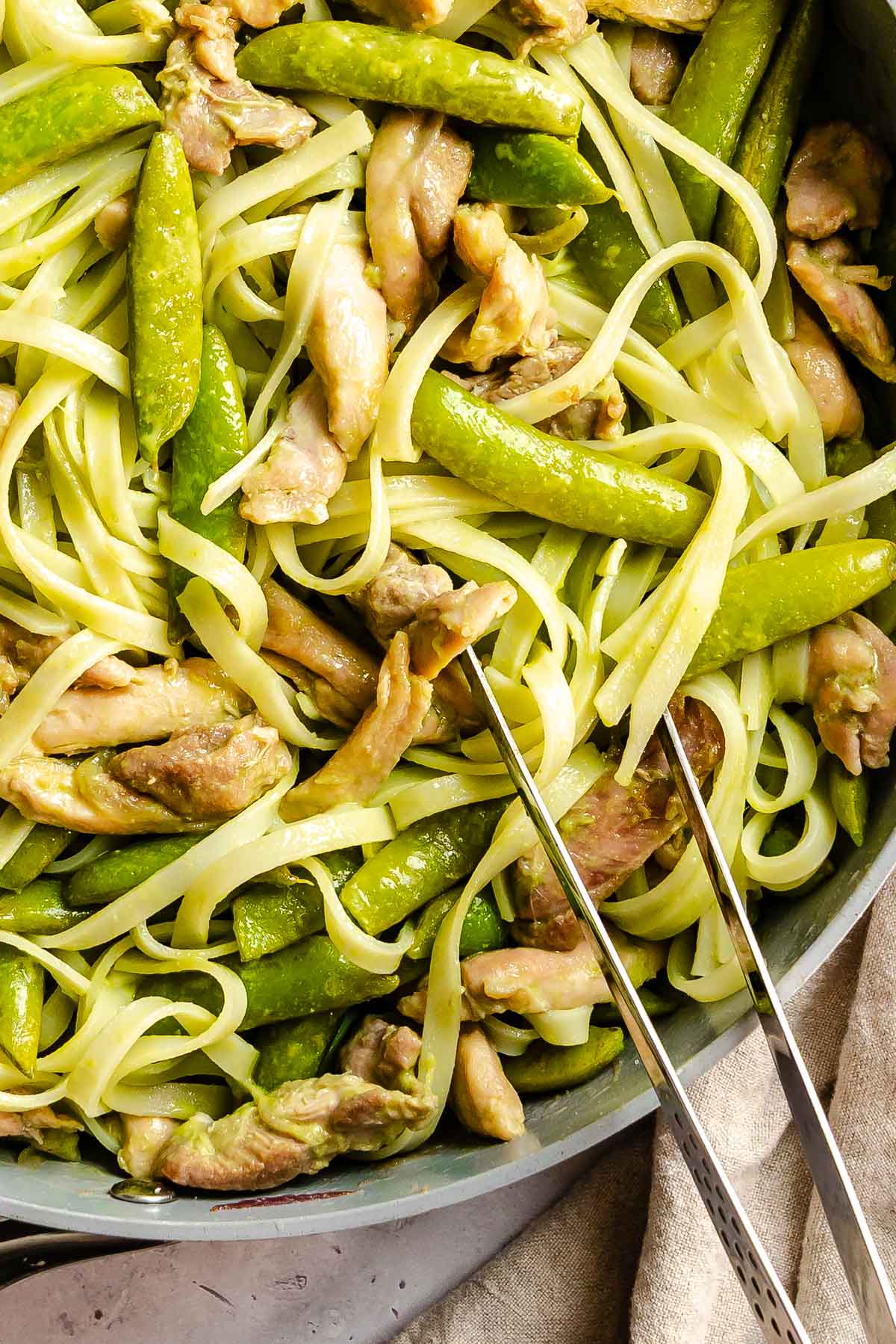 Rice noodles with green vegetables and chicken in a pan with tongs, ready to serve.