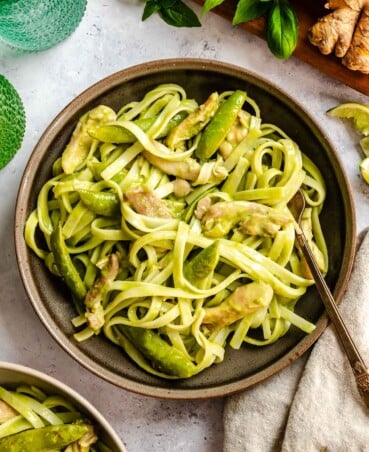 A bowl of pasta with chicken and snap peas in a creamy sauce, with a fork and fresh ginger nearby.