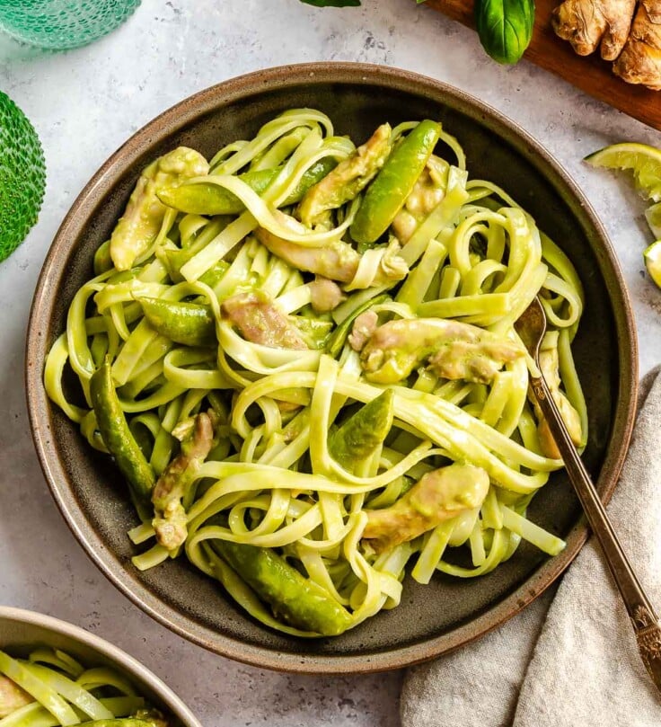 A bowl of pasta with chicken and snap peas in a creamy sauce, with a fork and fresh ginger nearby.