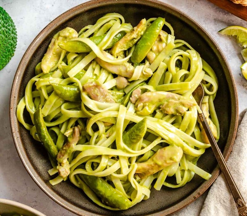 A bowl of pasta with chicken and snap peas in a creamy sauce, with a fork and fresh ginger nearby.