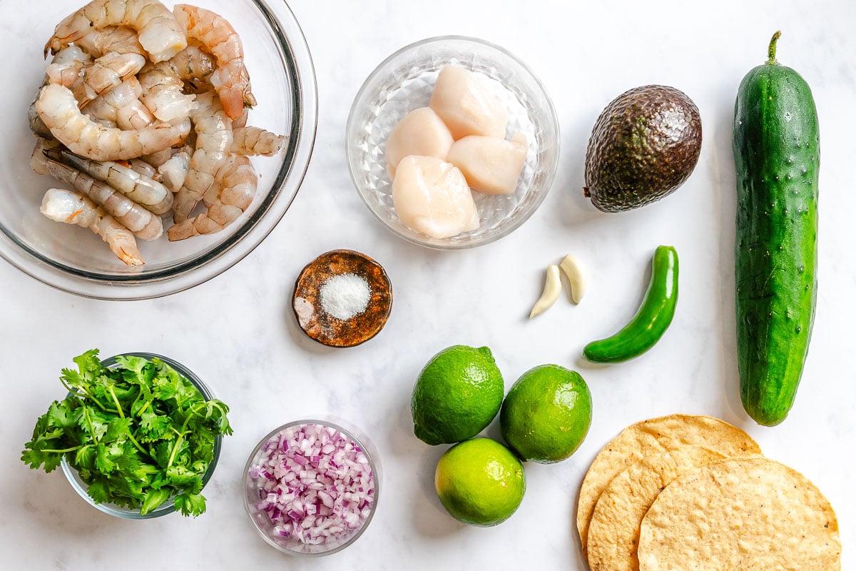 Raw shrimp, scallops, avocado, cucumber, limes, tostadas, cilantro, red onion, salt, garlic, serano pepper set out on a white counter.