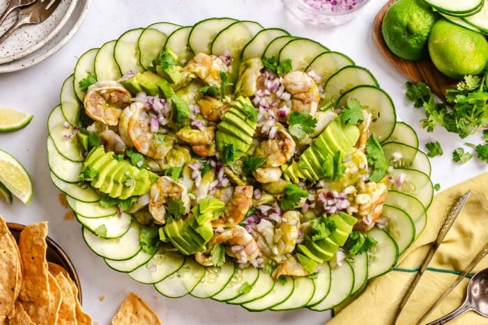Raw shrimp and scallop in a vibrant green sauce on a large platter lined with thinly sliced cucumnbers and topped with minced recipe onion, sliced avocado and cilantro.