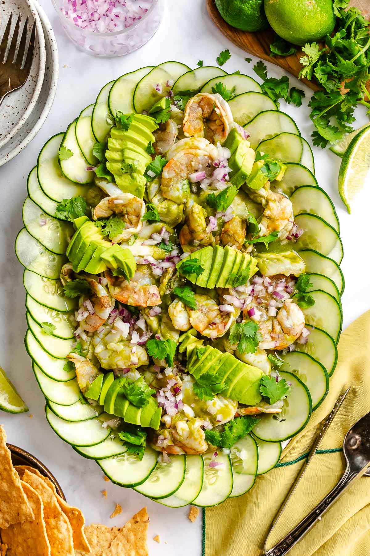 Raw shrimp and scallop in a vibrant green sauce on a large platter lined with thinly sliced cucumnbers and topped with minced recipe onion, sliced avocado and cilantro.