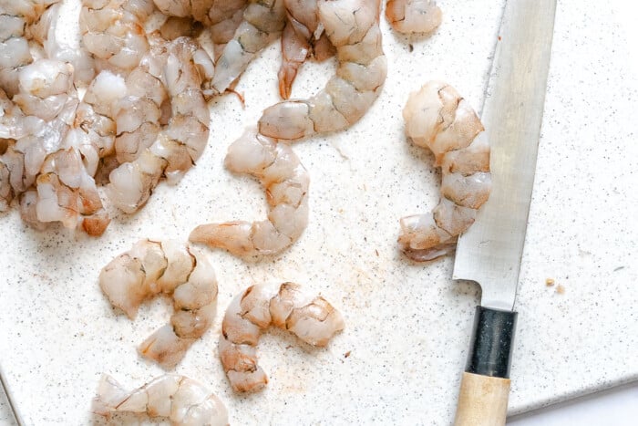 Raw shrimp and a knife on a white cutting board, ready for preparation.