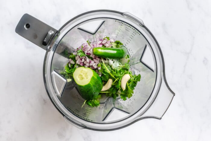Chopped red onion, cilantro, garlic, cucumber, and serano in a blender on a white marble surface.