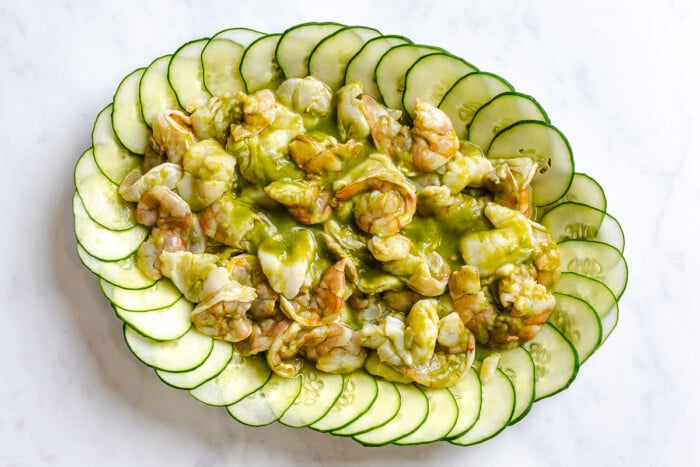 Shrimp and scallop aguachile on a large platter lined with thinly sliced cucumnbers and topped with minced recipe onion, sliced avocado and cilantro.