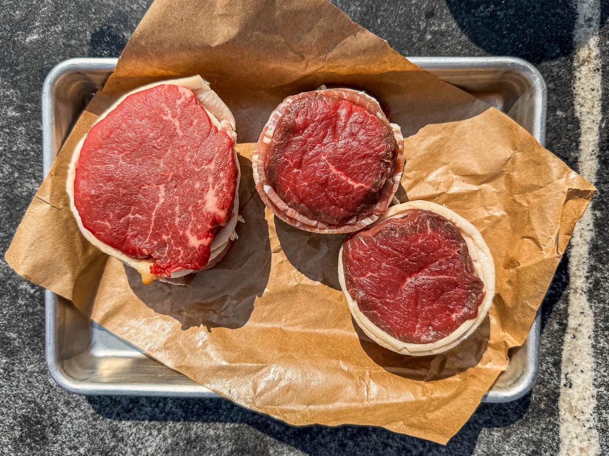 Bacon wrapped filet steaks on a parchment-lined baking sheet.