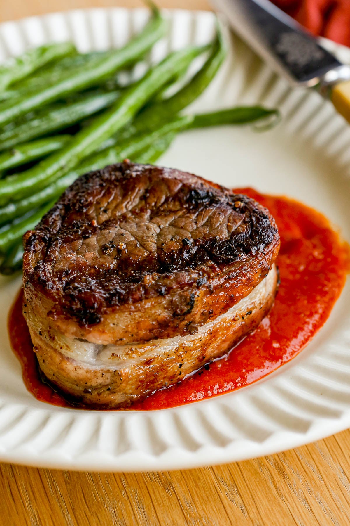 Bacon-wrapped filet mignon on red sauce with green beans on a white plate.