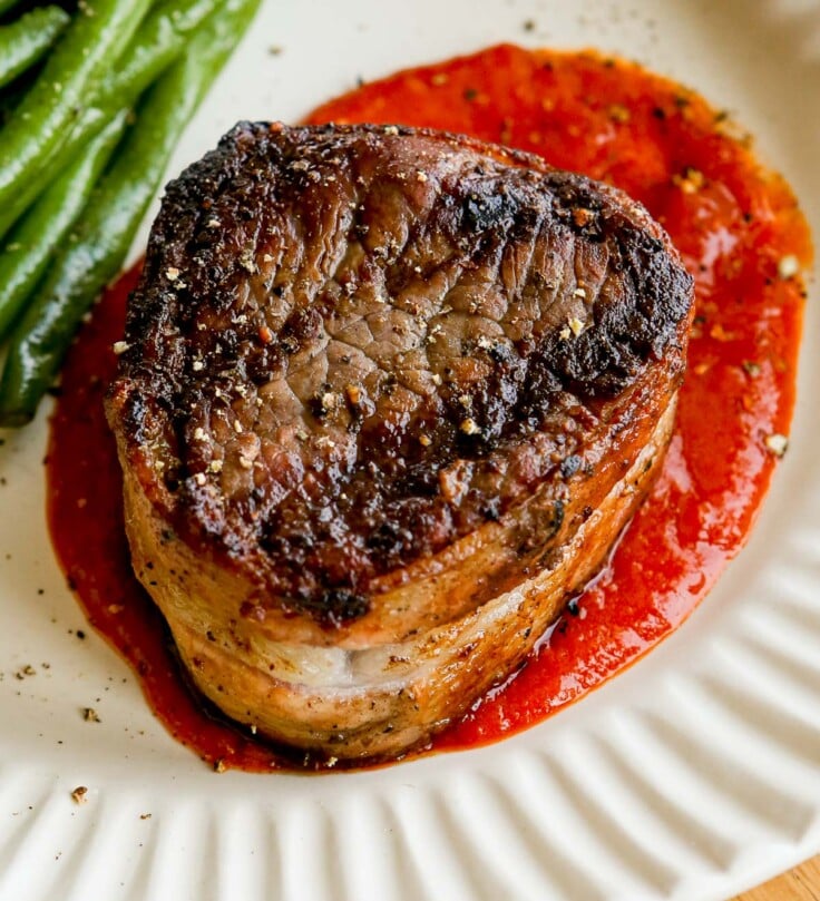 Bacon-wrapped filet mignon on red sauce with green beans on a white plate.