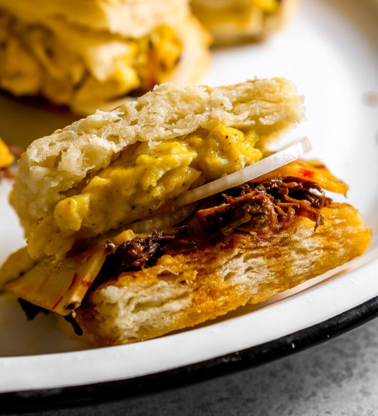 Close-up of a breakfast biscuit sandwich with scrambled eggs, cheese, onion, and shredded beef on a white plate.