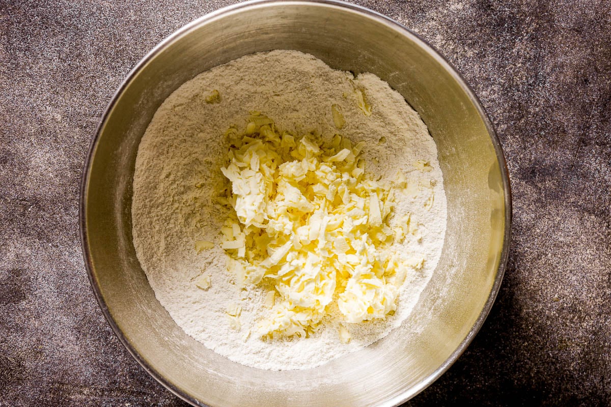 A metal bowl with flour and grated butter, ready to be mixed on a dark countertop.