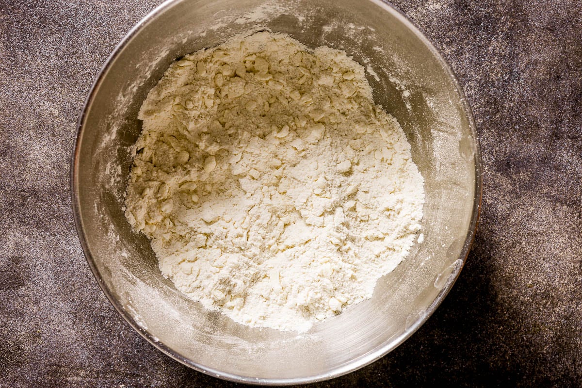 Metal bowl with flour and chunks of butter, ready for mixing on a dark countertop.