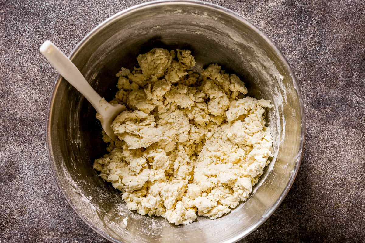 A metal bowl with dough being mixed by a white spatula, on a dark textured surface.