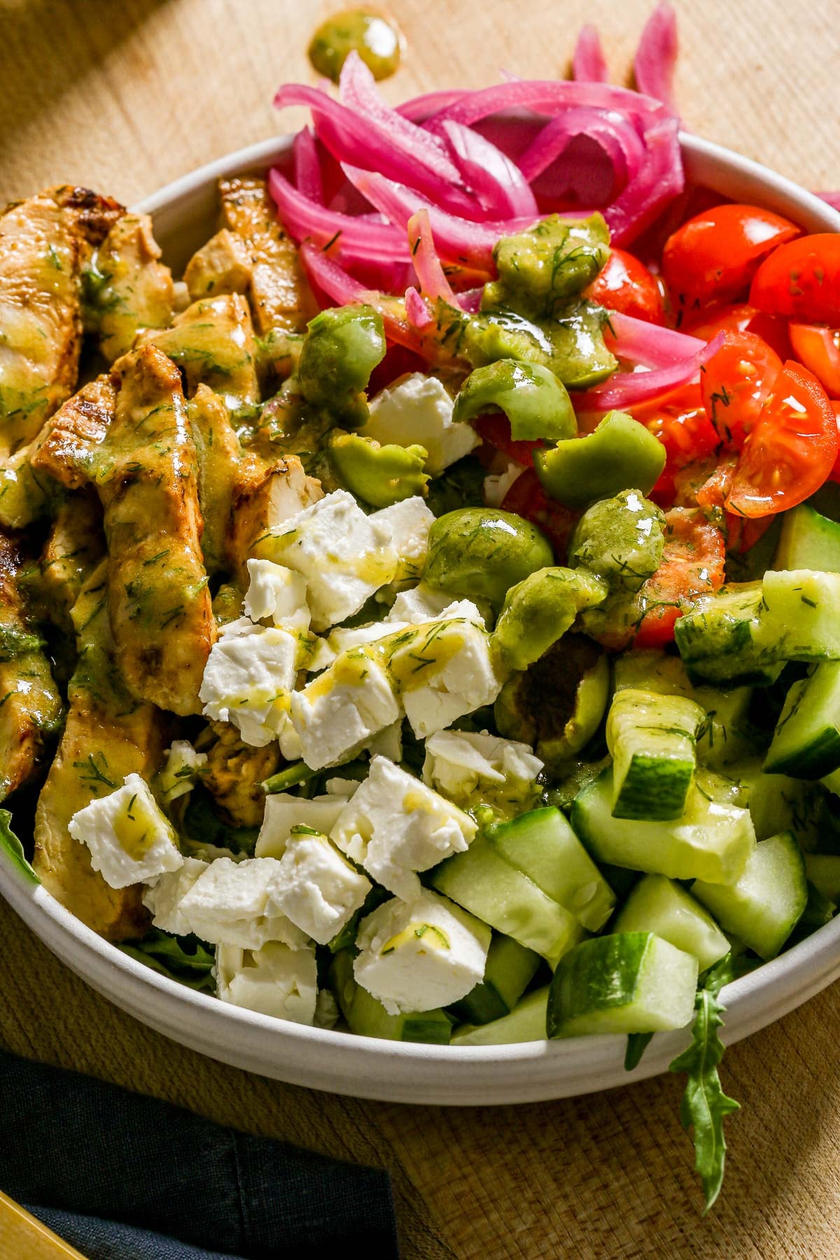 A bowl with cooked chicken, feta, cucumber, tomatoes, olives, pickled onions, and green dressing.