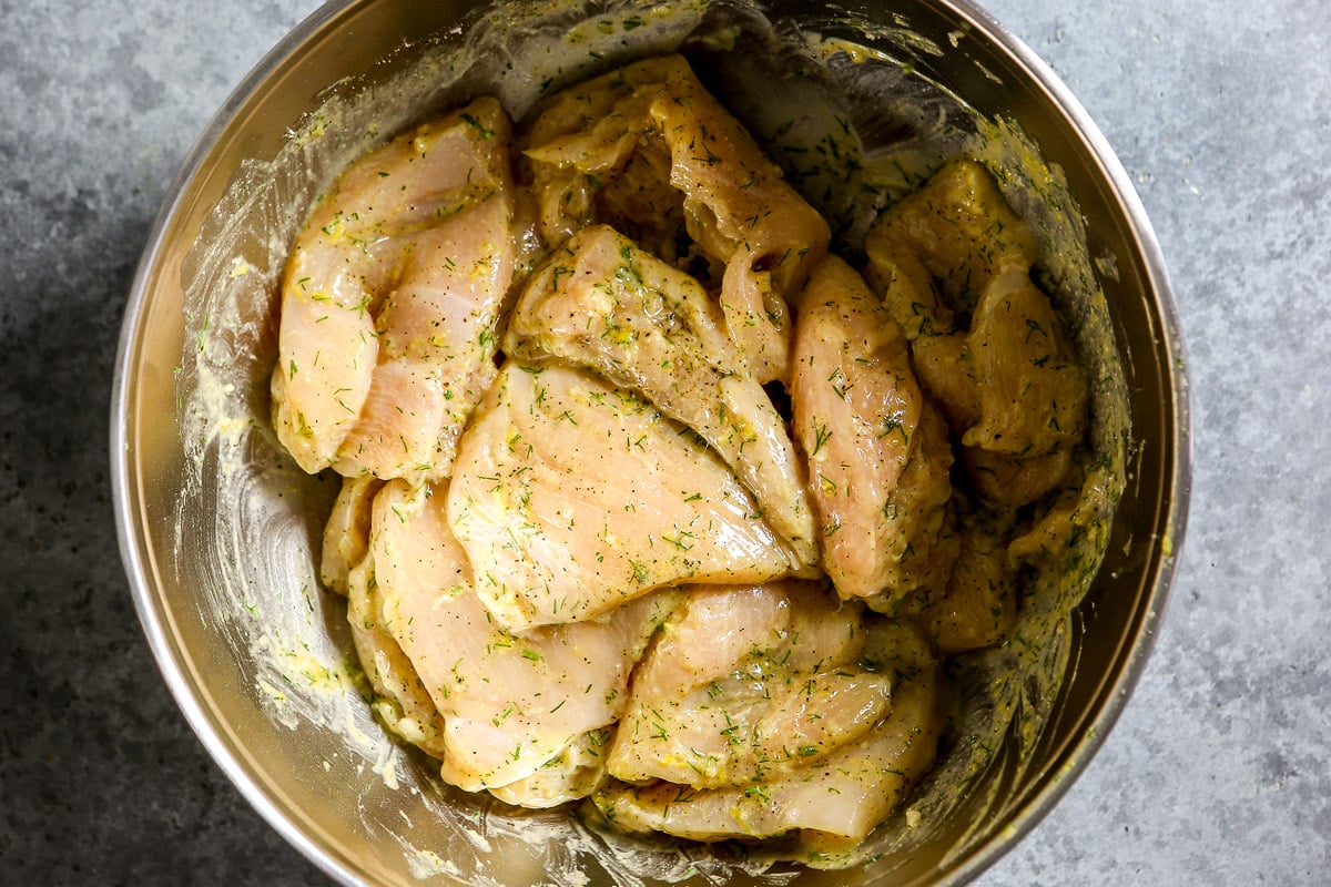 Chicken breasts marinating in a lemon and dill sauce in a large metal bowl.
