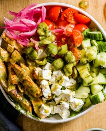 A bowl with browned chicken breast, feta, cucumber, tomatoes, olives, pickled onions, and green dressing.