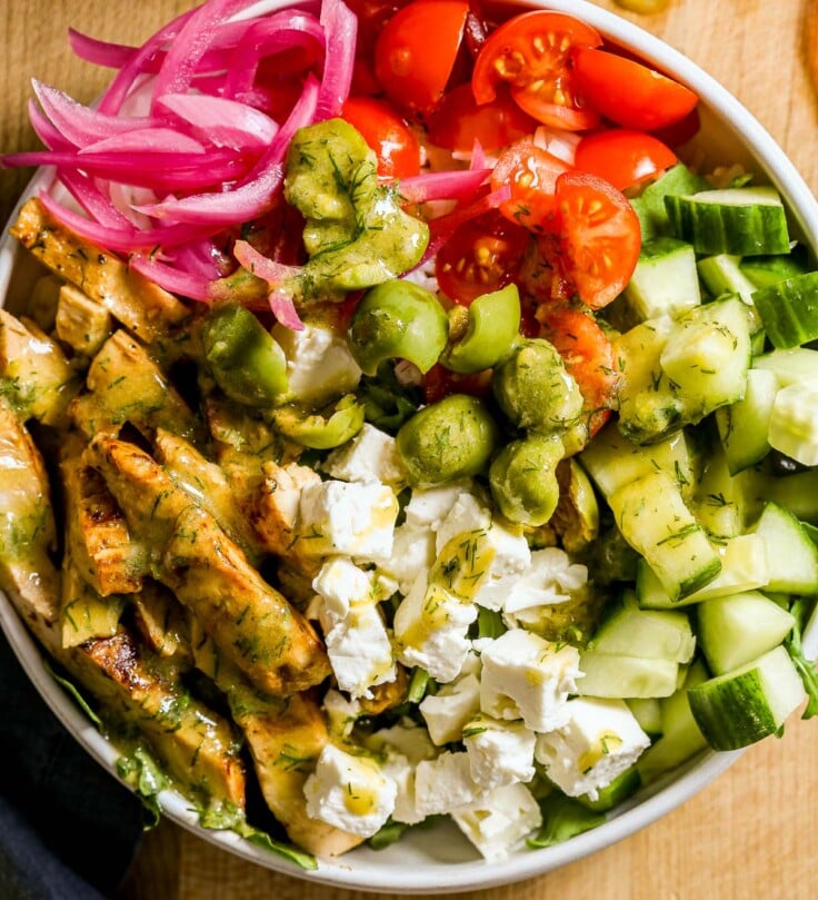 A bowl with browned chicken breast, feta, cucumber, tomatoes, olives, pickled onions, and green dressing.
