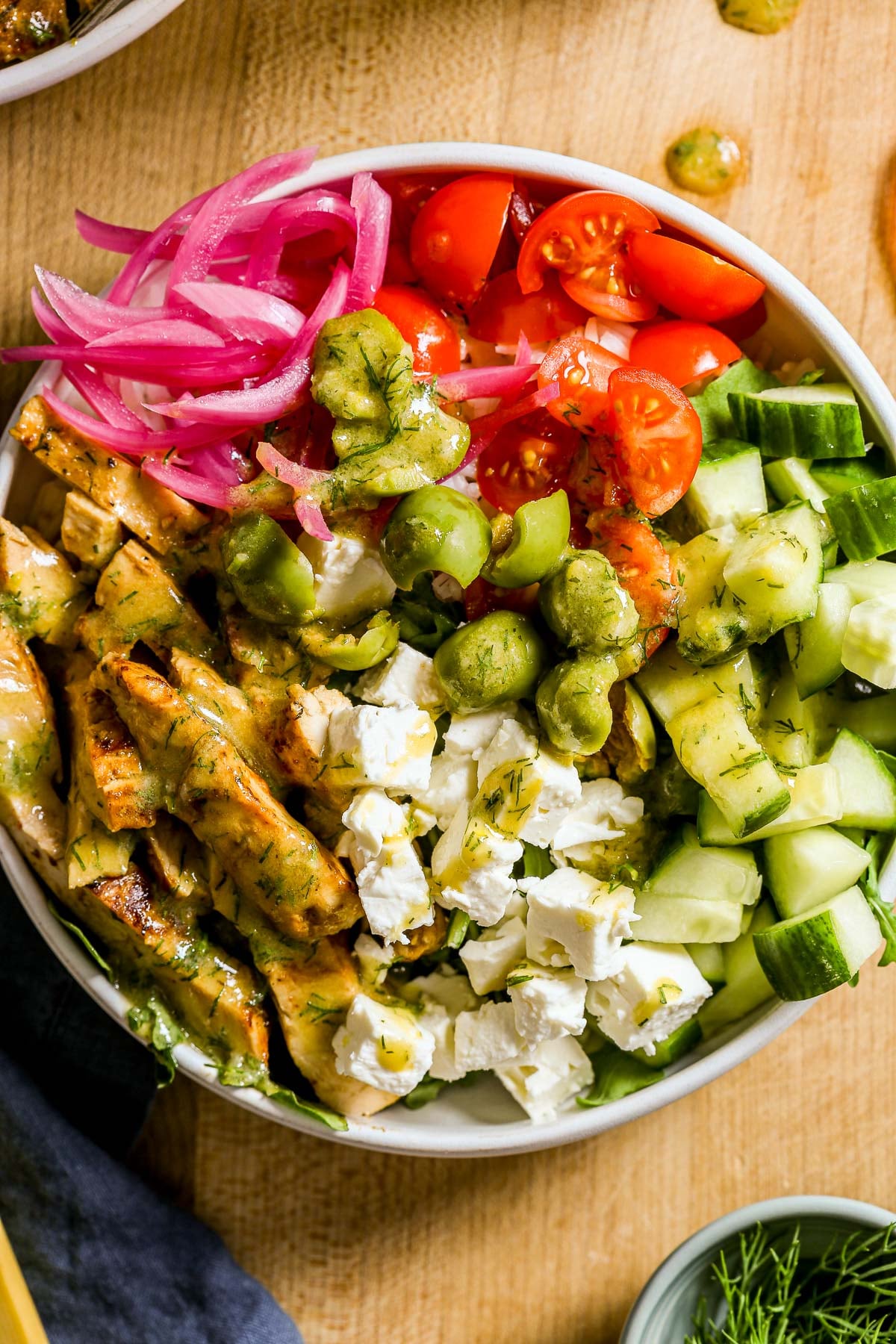 A bowl with browned chicken breast, feta, cucumber, tomatoes, olives, pickled onions, and green dressing.