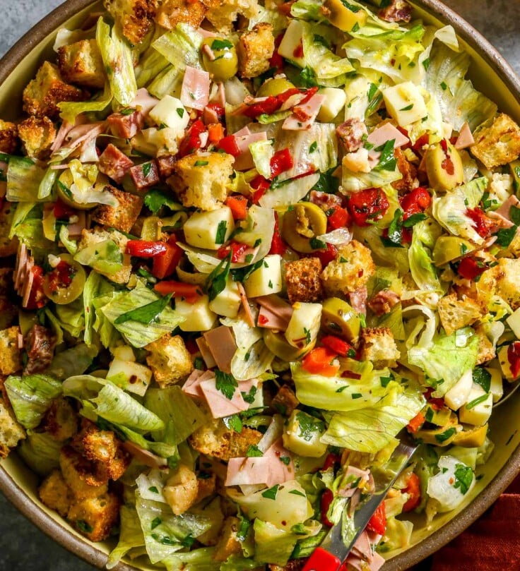 Chopped salad with lettuce, ham, cheese, olives, peppers, croutons, and parsley in a large bowl with a red spoon.