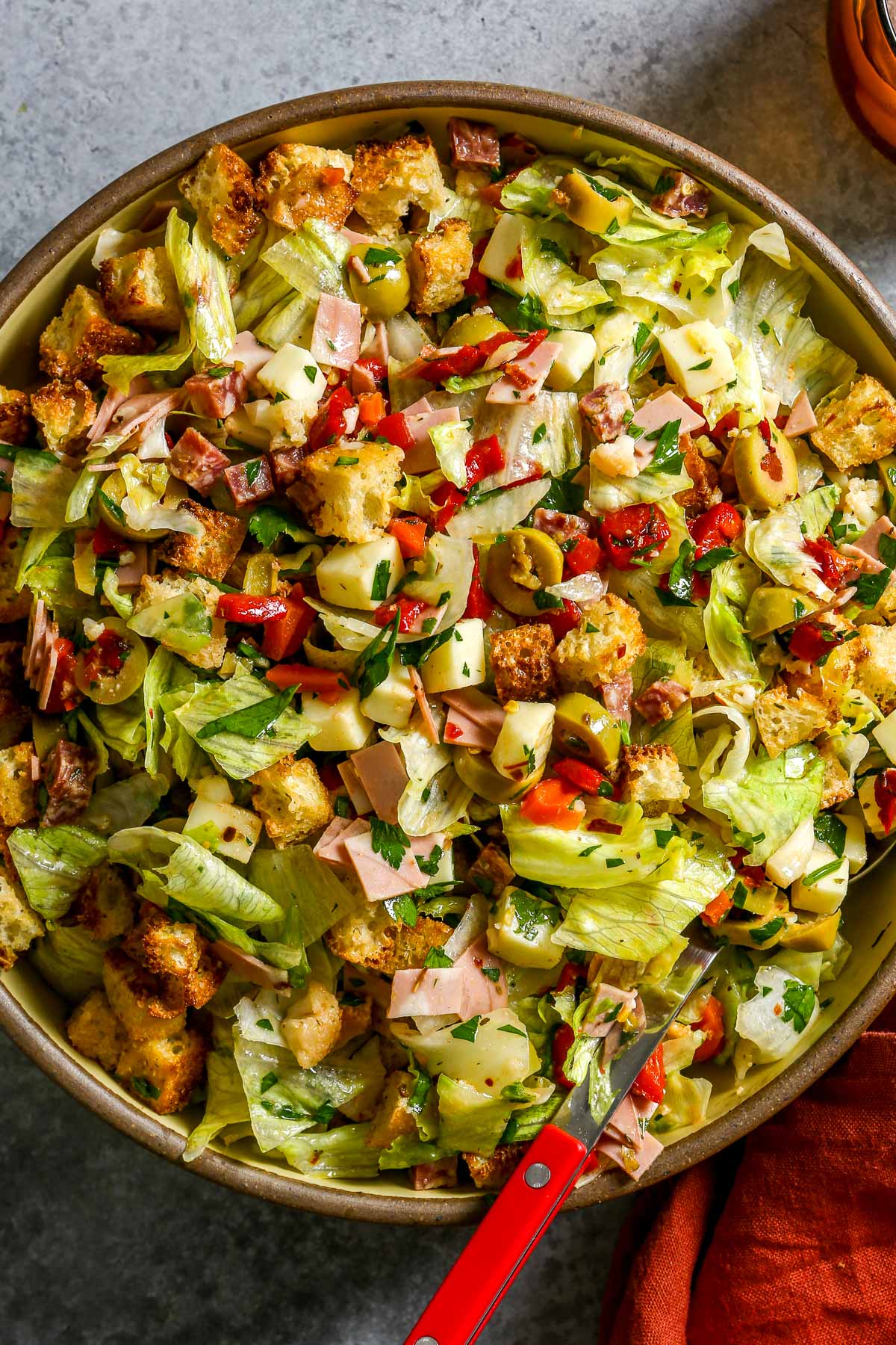 Chopped salad with lettuce, ham, cheese, olives, peppers, croutons, and parsley in a large bowl with a red spoon.