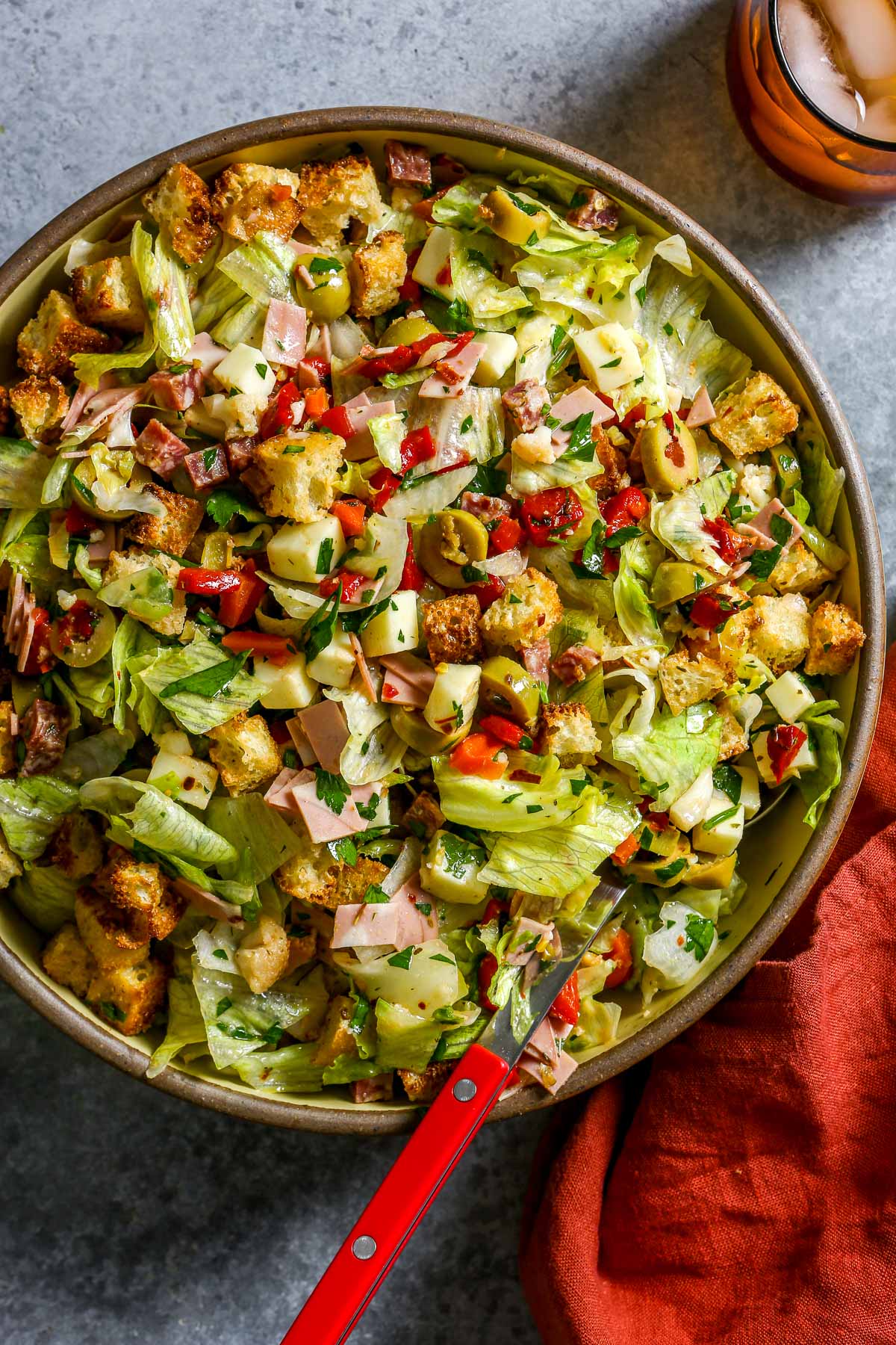 Chopped salad with lettuce, ham, cheese, olives, peppers, croutons, and parsley in a large bowl with a red spoon.