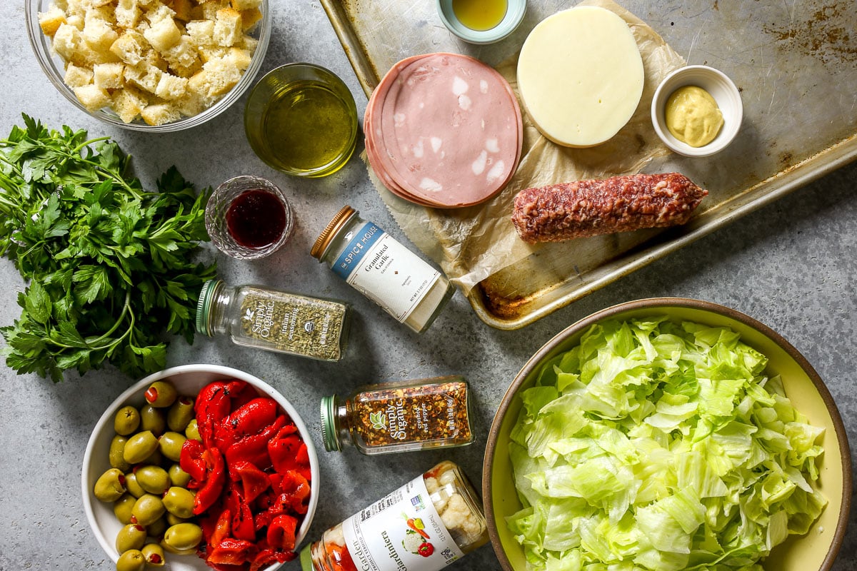 Olives, roasted red bell peppers, fresh parsley, cubes of bread, olive oil and vinegar, spices, giardiniera, mortadella, provolone, salami, Dijon, honey, and lettuce all set out on a counter ready to be turned into a salad.