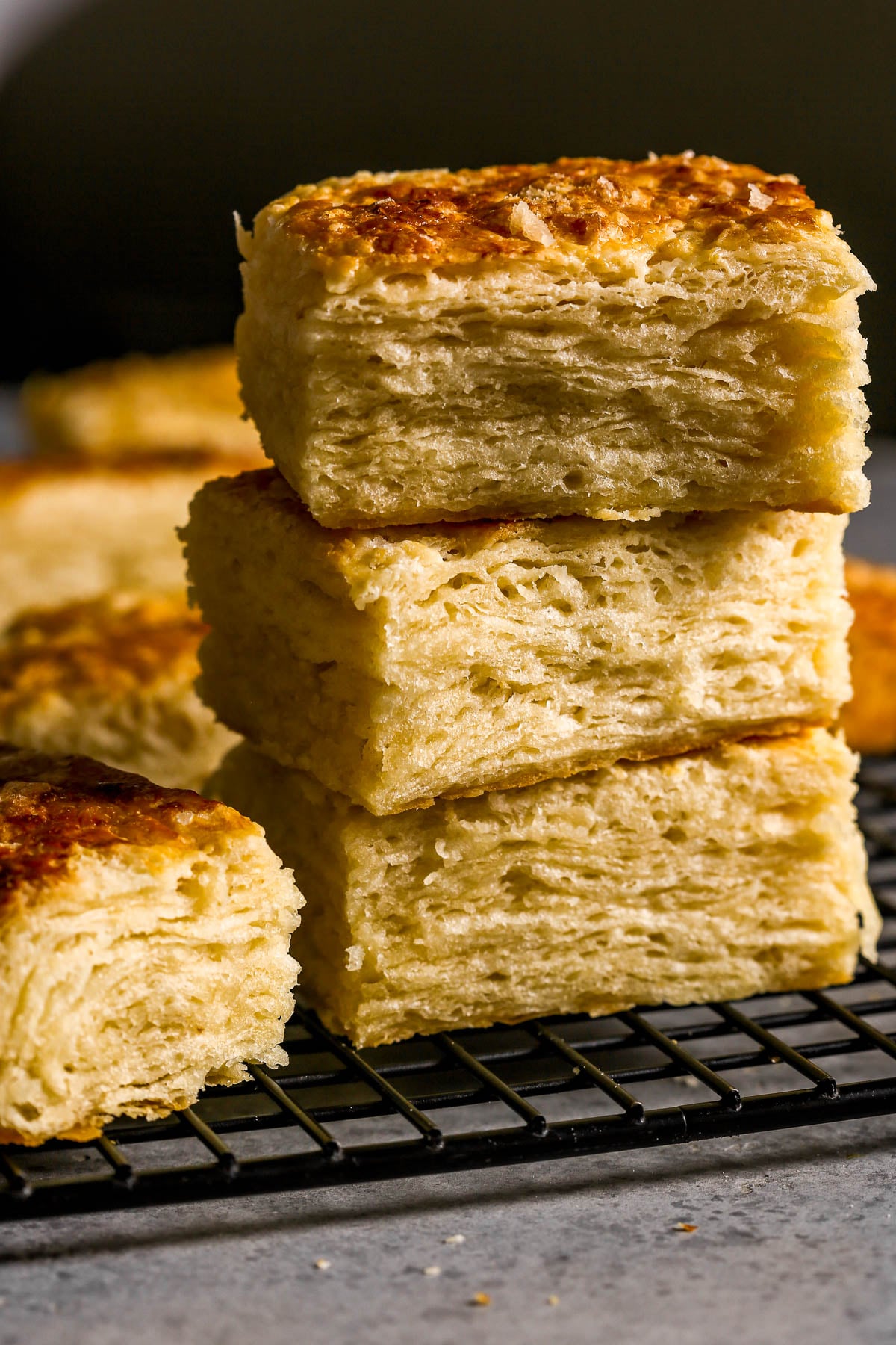 Three flaky, golden-brown biscuits are stacked on a black cooling rack with more biscuits in the background.