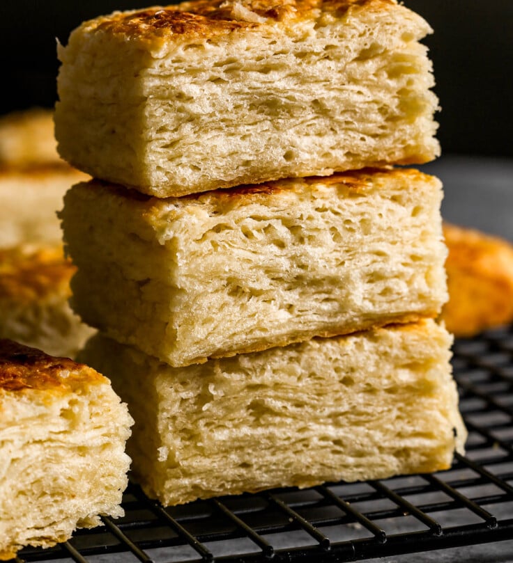Three flaky, golden-brown buttermilk biscuits are stacked on a black cooling rack with more biscuits in the background.