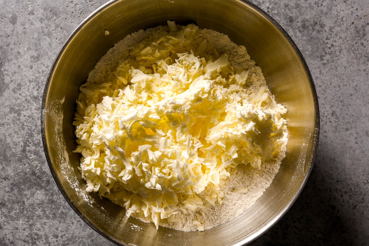 A metal bowl with flour and a pile of shredded butter on a gray countertop.